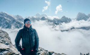Trevor smiling at the camera. In the background there are large, snowy mountains. This was taken on his trip to Nepal.