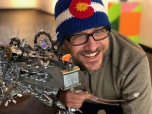Trevor smiling at the camera with a piece of art that he made. He is wearing a knitted beanie designed as the Colorado flag. His sculpture is made out of metal.