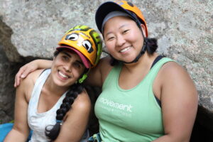 Jess and Esha both wearing climbing helmets sitting together in front of a rock face smiling. Jess has her arm around Esha's shoulder.