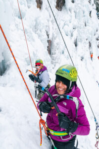 Esha ice climbing in the Ouray Ice Park. She is smiling at the camera and has her ice axes crossed in front of her. Photo by Will Strathmann.