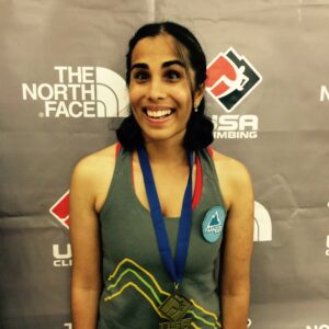 Esha smiling wearing a medal from the 2015 Paranationals. She is standing in front of a USA Climbing and The North Face backdrop.