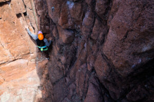 Jess mid-climb in Staunton SP. The photo is taken from above, looking down towards Jess on the wall. She has her left hand high and is looking up. She is wearing a bright orange helmet.