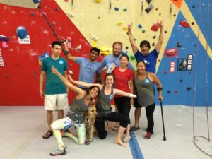 Jess standing with a crew of Paradox climbers in front of an indoor climbing wall. Taken from a Paraclimbing competition. Everyone is either wearing Paradox t-shirts or has a Paradox patch on their shirt. All 8 of them (plus one dog) are looking at the camera and smiling. 