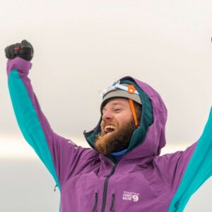 Tanner Jones on top of Volcan Cotopaxi. He has a big smile and his arms outstretched in the air. He is wearing a bright purple and blue coat with a grey helmet on.