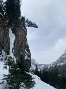 Erich climbing a frozen waterfall. The ground is snowy and there are rocky, snowy cliffs on the right.