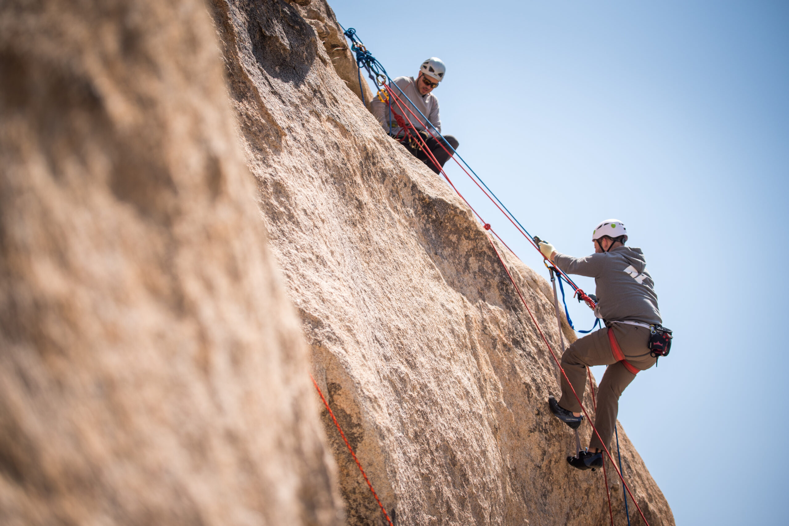 A side view of a climber using an aiding system in Joshua Tree. They are stepping up a ladder aider. There is a guide at the top of the route above them looking down towards them.