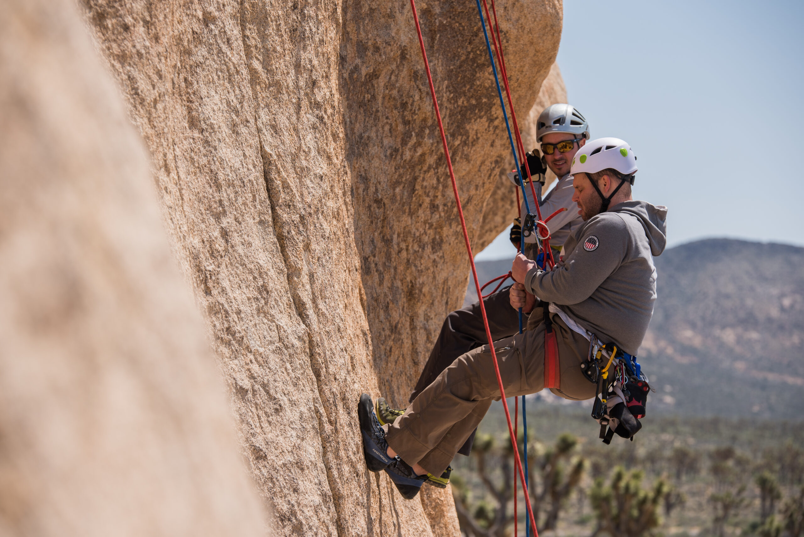 A climber practing rappelling with the help of a guide who is side by side with him. There is a mountain and Joshua Trees behind them.