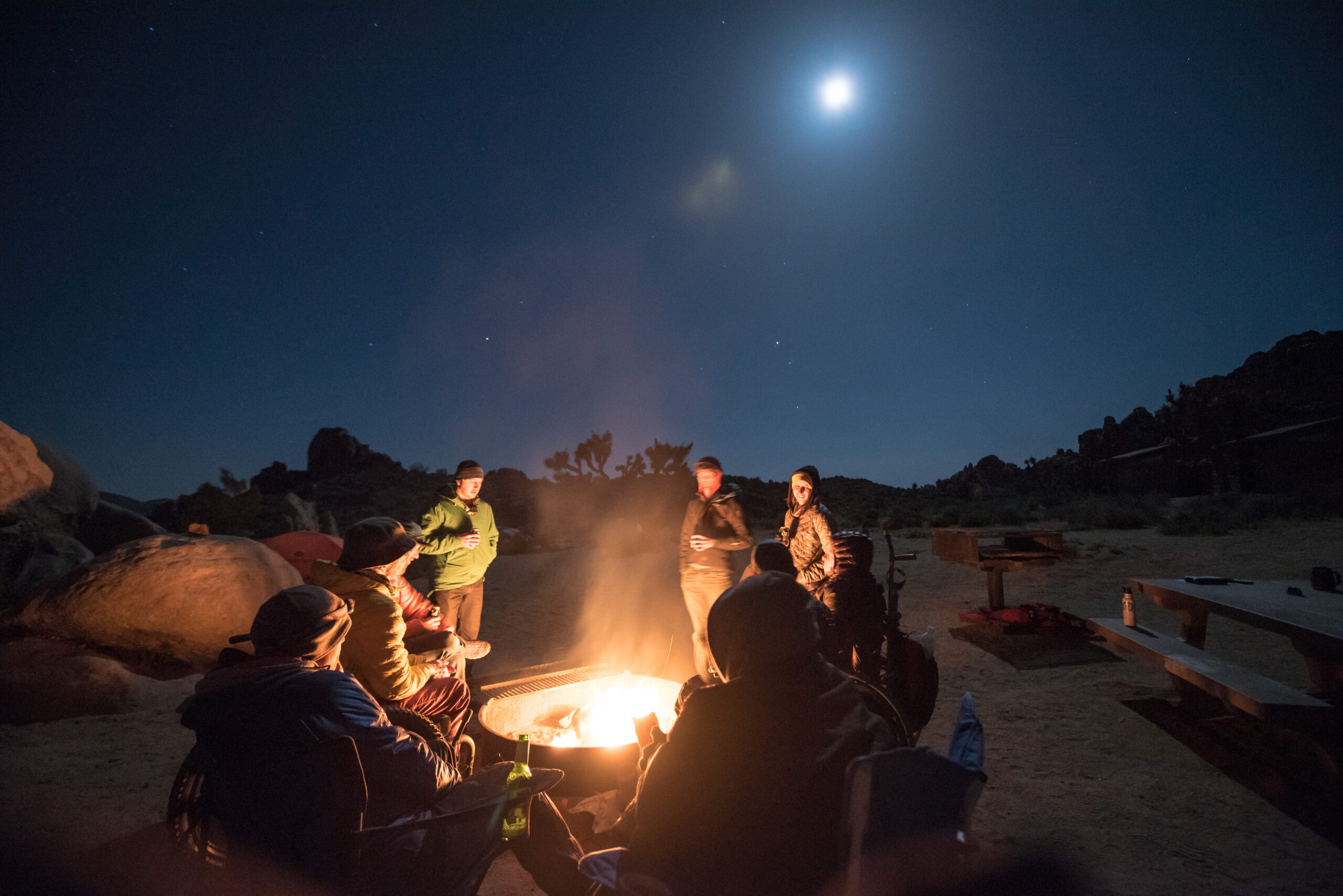 A night picture of a group of climbers sitting around a fire in Joshua Tree. The sky is dark and you can see some stars in the sky. Everyone is in jackets and hats.