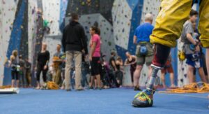 An image of a prosthetic limb wearing a climbing shoe. In the background there are many people gathered in a climbing gym. Some are climbing and others are standing on the ground.