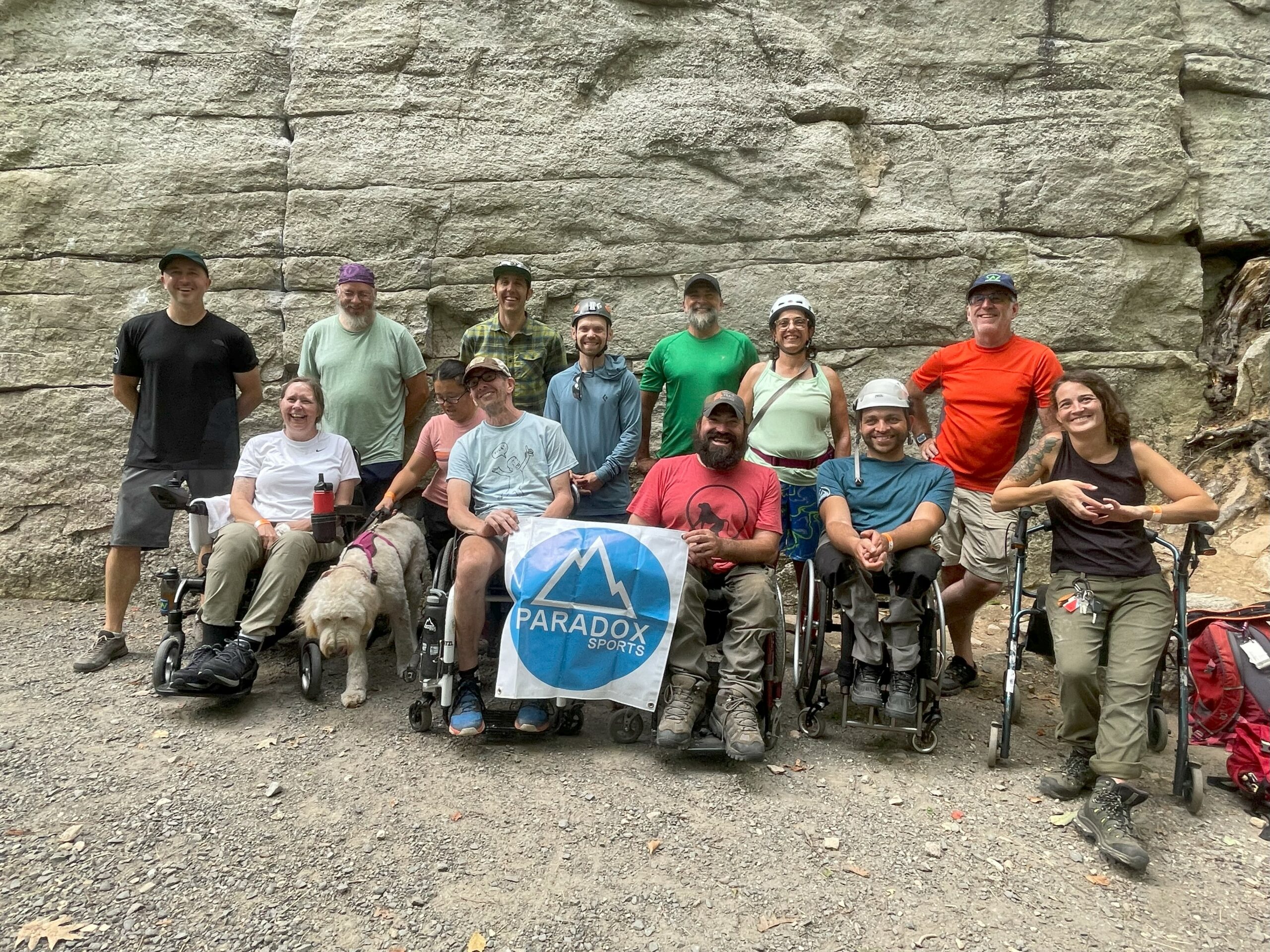 A group shot at the Gunks. The group is smiling at the camera and holding a Paradox Sports banner. They are standing in front of a rock wall.